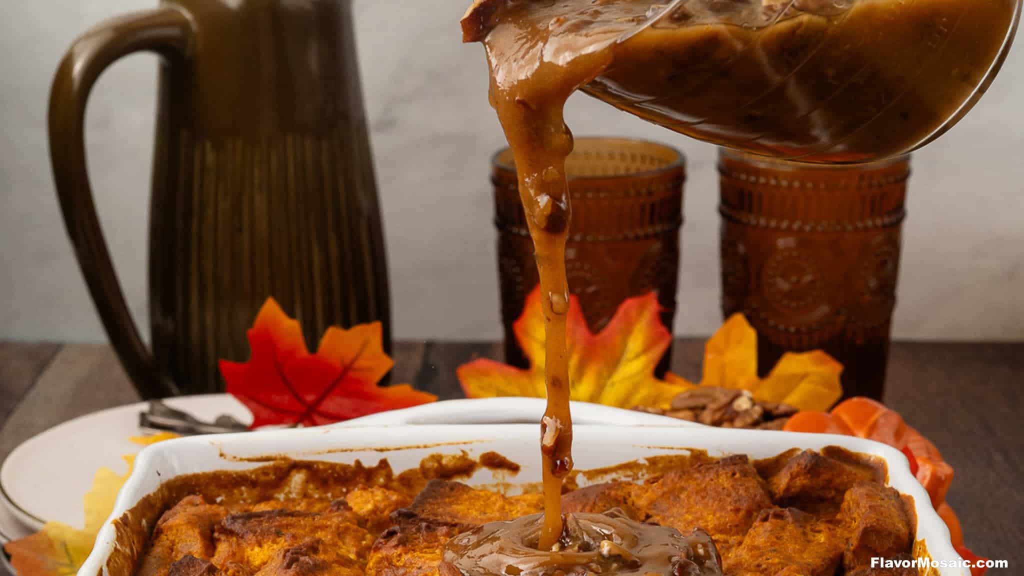 Caramel sauce is being poured from a glass pitcher onto Pumpkin Praline Bread Pudding in a white dish. Decorative autumn leaves, pecans, two brown glasses, and a ceramic jug are in the background, creating a cozy fall atmosphere.