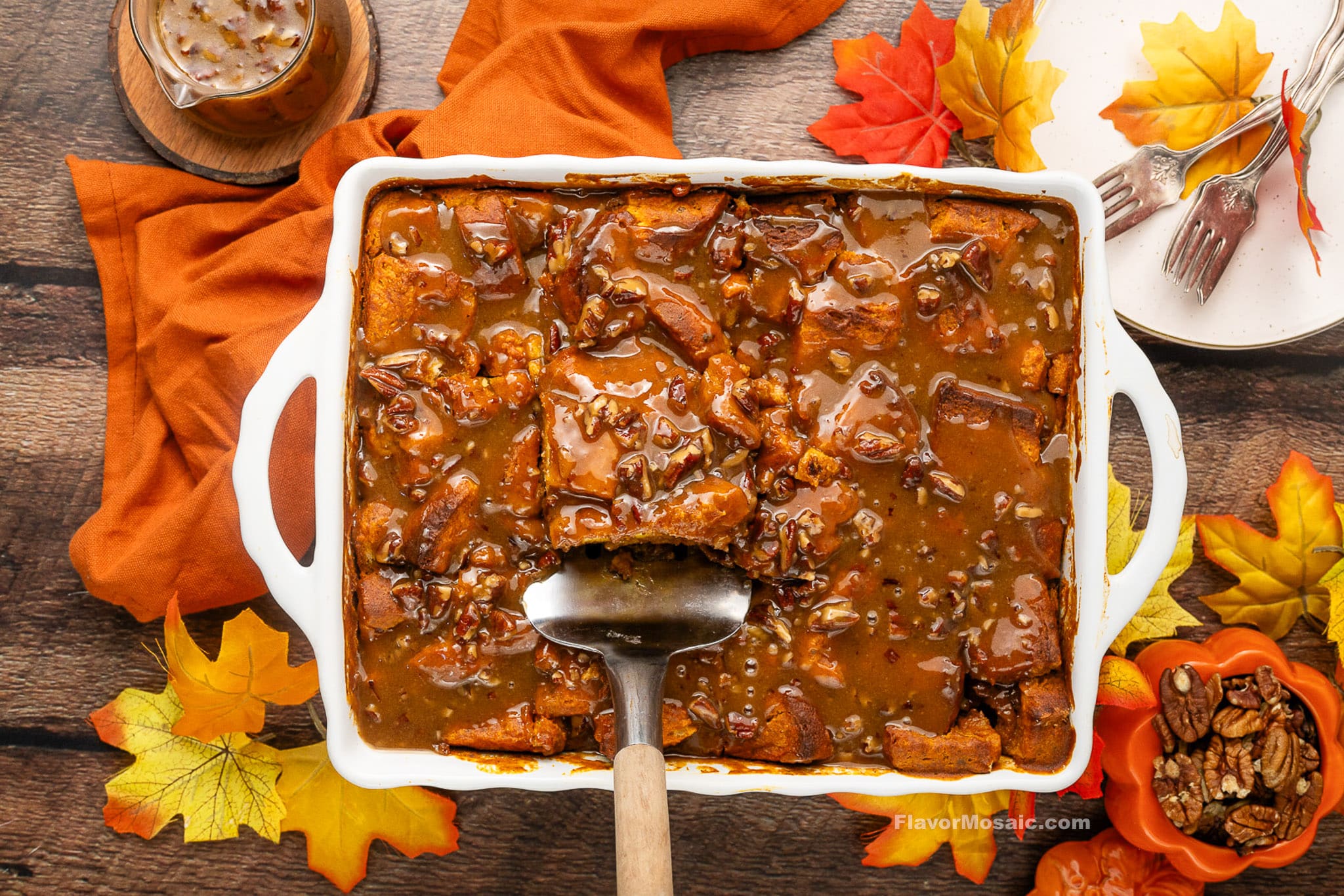 A white baking dish filled with sweet potato casserole topped with pecans and brown sauce, reminiscent of a Pumpkin Praline Bread Pudding, is surrounded by autumn leaves, an orange napkin, a drink, forks, and a small bowl of pecans on a wooden table.