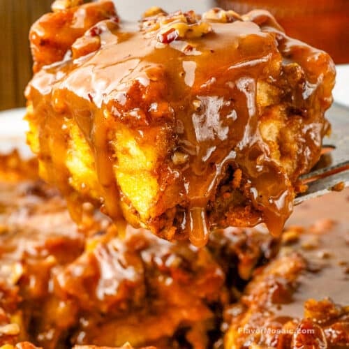 A gooey slice of Pumpkin Praline Bread Pudding covered in caramel sauce is lifted from a dish, showing its moist texture and pecan topping.