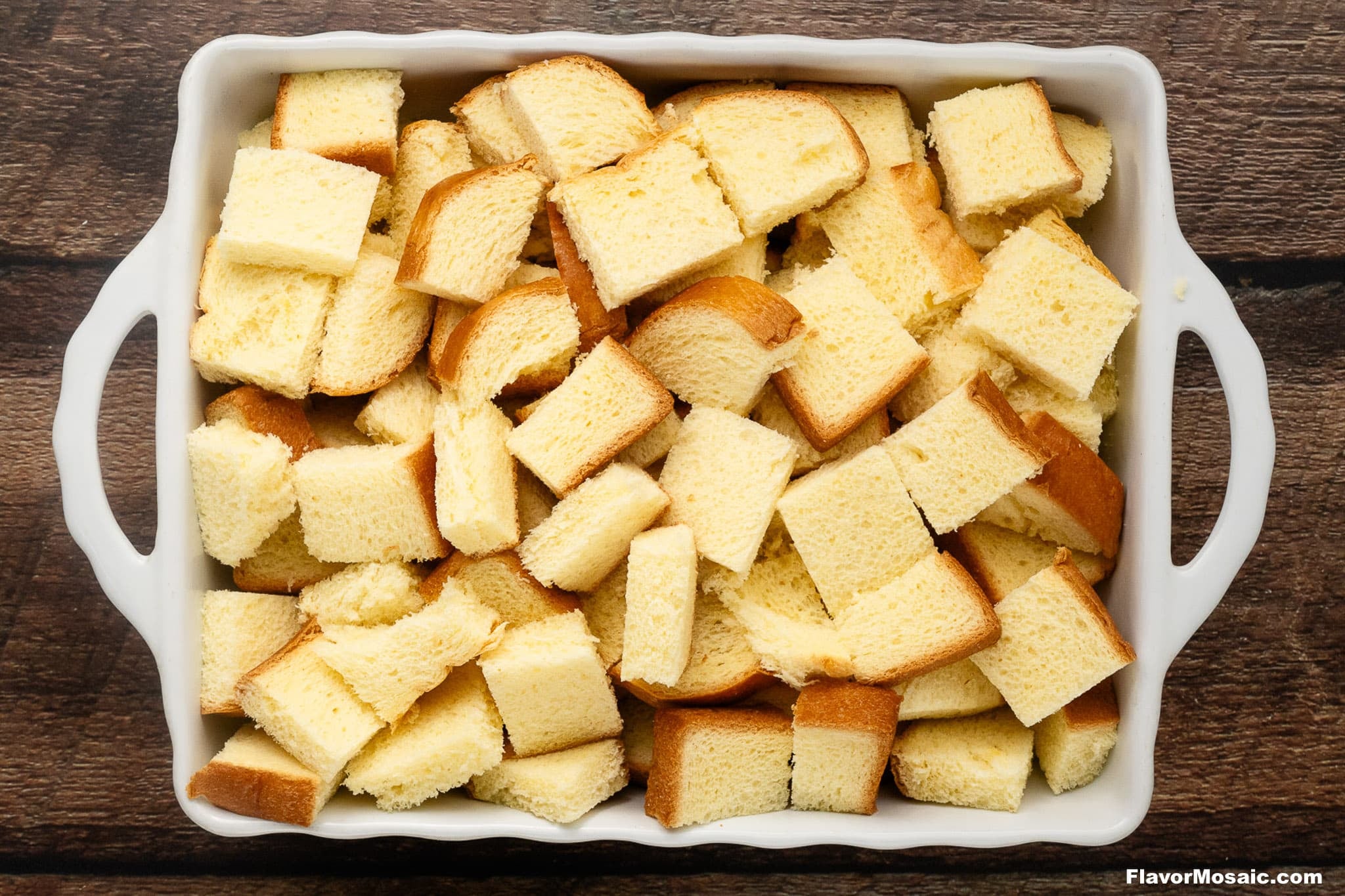 Cubes of bread are arranged in a white rectangular baking dish, ready to transform into a delicious Pumpkin Praline Bread Pudding. The dish rests on a wooden surface, awaiting its time in the oven.