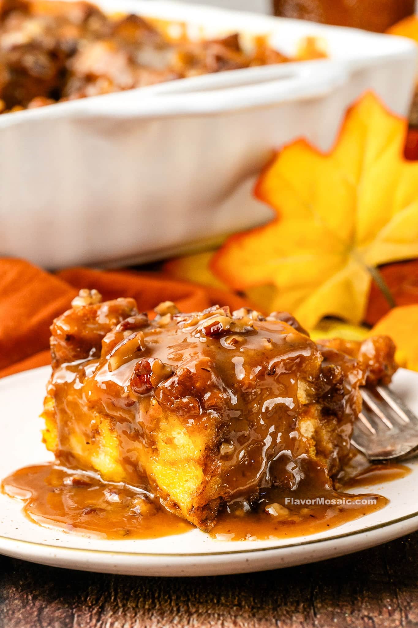 A slice of Pumpkin Praline Bread Pudding topped with caramel pecan sauce sits on a white plate, with autumn leaves and a baking dish in the background.