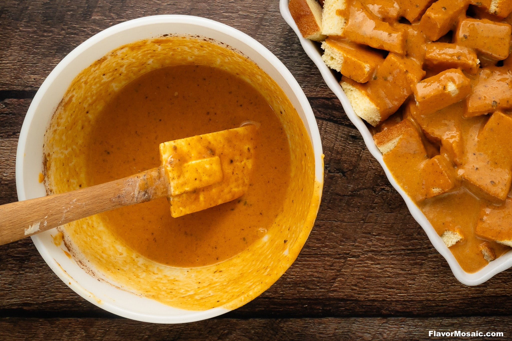 Overhead shot of a mixing bowl containing a spiced, pumpkin-colored sauce with a wooden spatula inside, next to a baking dish filled with cubed bread coated in the same sauce-perfect for creating Pumpkin Praline Bread Pudding-all set on a dark rustic wooden background.