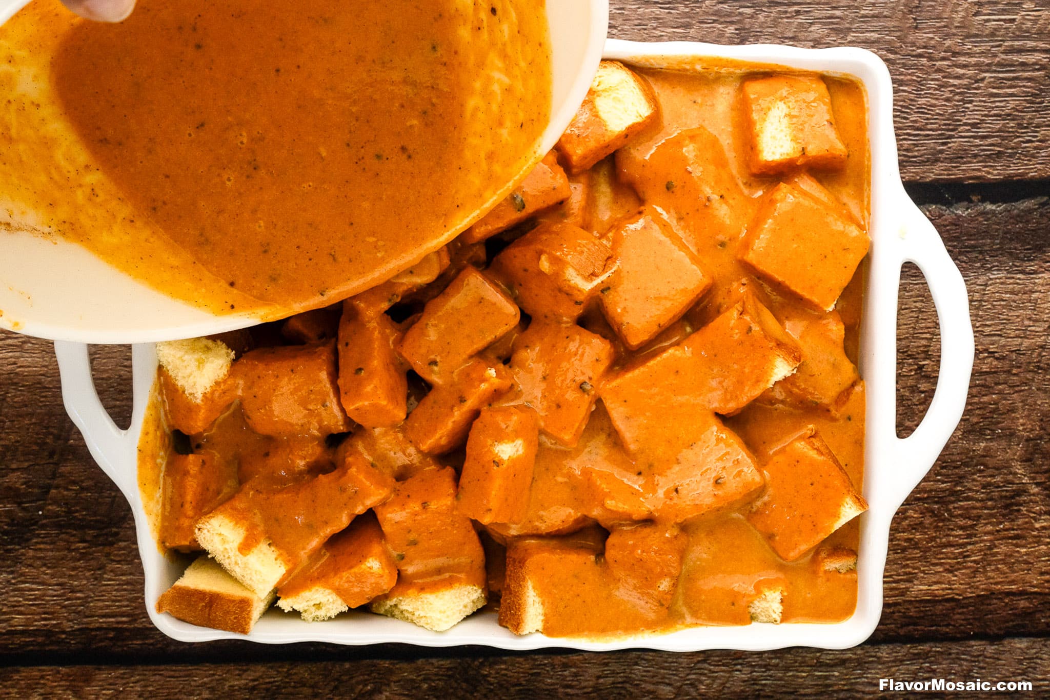 Cubed bread is covered with a luscious orange sauce in a white baking dish while more is being poured on top from a bowl, creating an inviting Pumpkin Praline Bread Pudding, all set on a wooden surface.