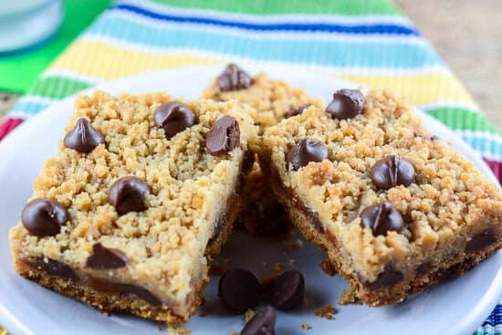 Three Chocolate Chip Caramel Crumb Bars are arranged on a white plate. The bars have a golden brown, streusel-like topping with chocolate chips, and are set on a colorful, striped cloth background.