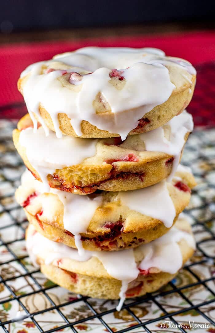 A stack of four baked Strawberry Donuts with visible pieces of fruit, topped with white icing, sits on a wire cooling rack as the icing drips down the sides.
