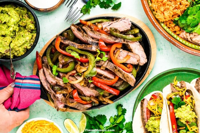 Overhead view of a sizzling platter of Tex-Mex Beef Fajitas with sautéed peppers and onions, surrounded by bowls of guacamole, Mexican rice, refried beans, shredded cheese, and tortillas ready to serve.