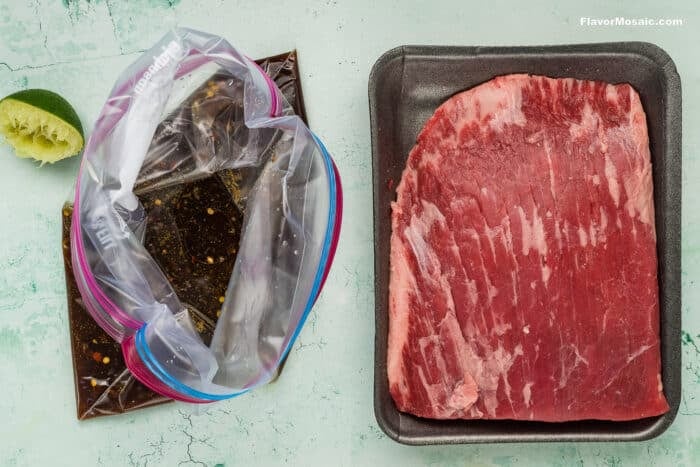 Raw flank steak on a black tray sits next to an open plastic bag of marinade, with a squeezed lime half above-perfect for making vibrant Tex-Mex Beef Fajitas on a light green, textured surface.