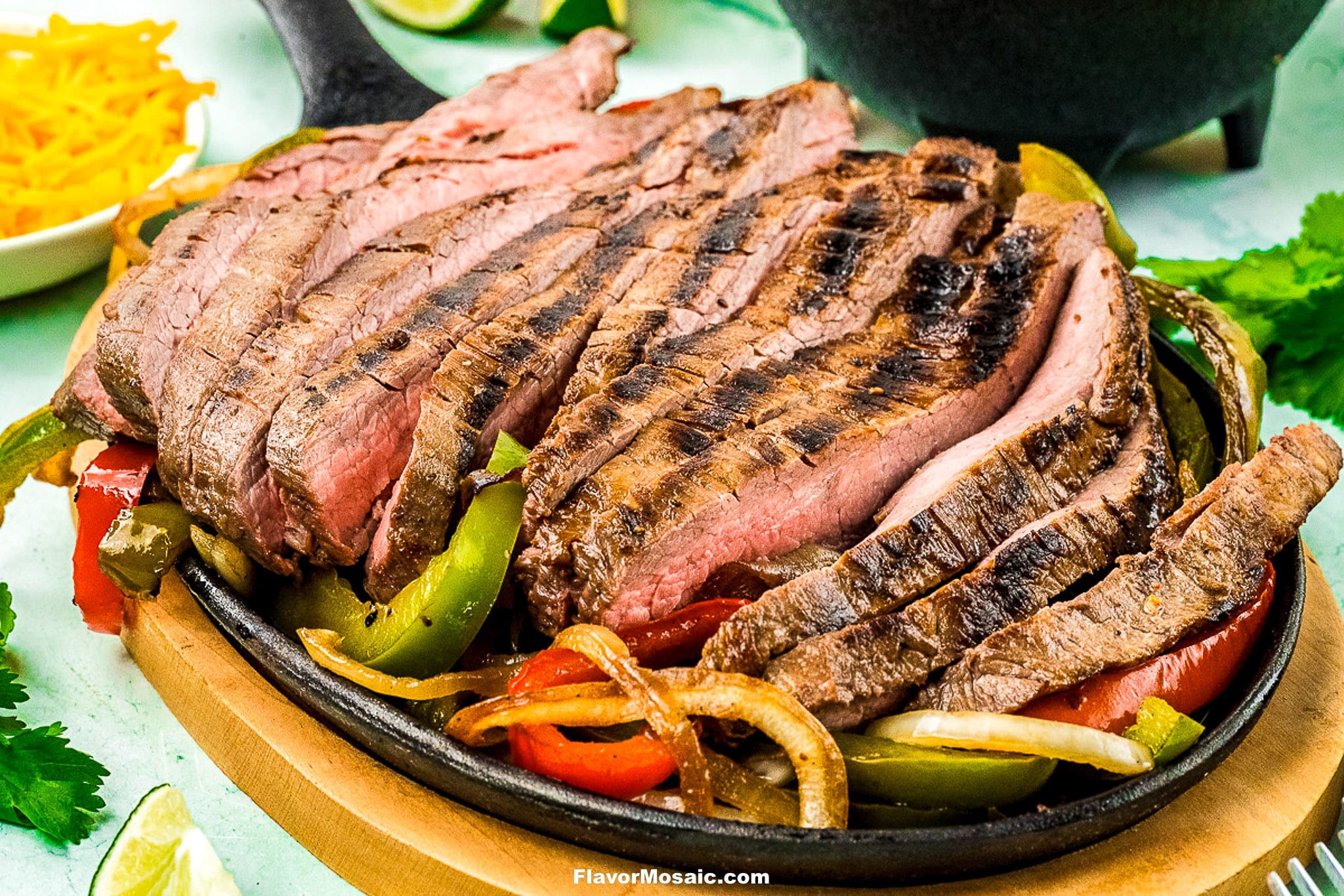 Sliced grilled steak, classic Tex-Mex Beef Fajitas style, served on a cast iron skillet with sautéed bell peppers and onions, garnished with cilantro. Shredded cheese and lime are visible in the background.