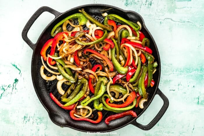 Sautéed red and green bell peppers with sliced onions in a black cast iron skillet on a light blue textured surface-perfect for serving alongside Tex-Mex Beef Fajitas.