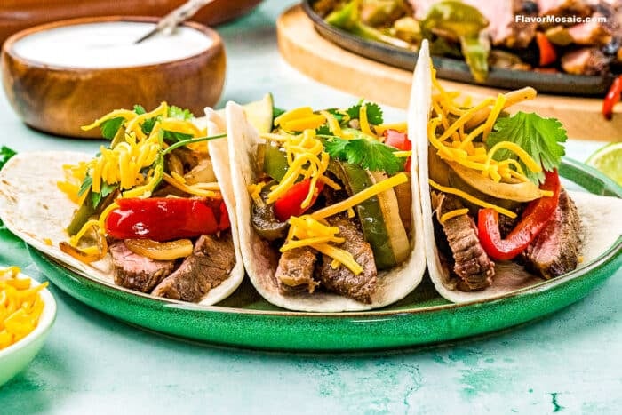 Three Tex-Mex Beef Fajitas tacos on flour tortillas, topped with sautéed peppers, onions, shredded cheddar cheese, and fresh cilantro, are arranged on a green plate. A bowl of sauce and extra shredded cheese are nearby.