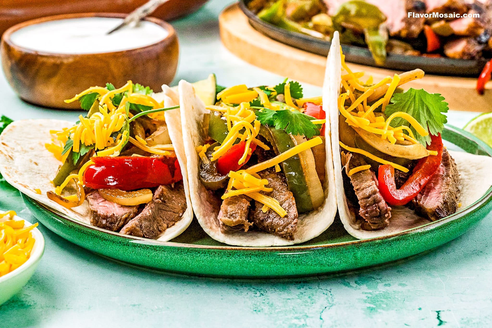 Three Tex-Mex Beef Fajitas tacos on flour tortillas, topped with sautéed peppers, onions, shredded cheddar cheese, and fresh cilantro, are arranged on a green plate. A bowl of sauce and extra shredded cheese are nearby.