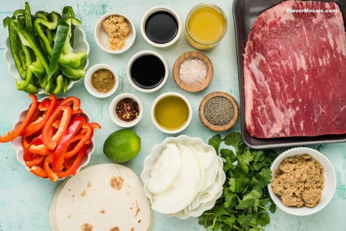 Ingredients for Tex-Mex Beef Fajitas laid out on a light blue surface: sliced green and red bell peppers, onions, cilantro, lime, tortillas, brown sugar, spices, soy sauce, garlic, oil, vinegar, salt, pepper, and raw steak.