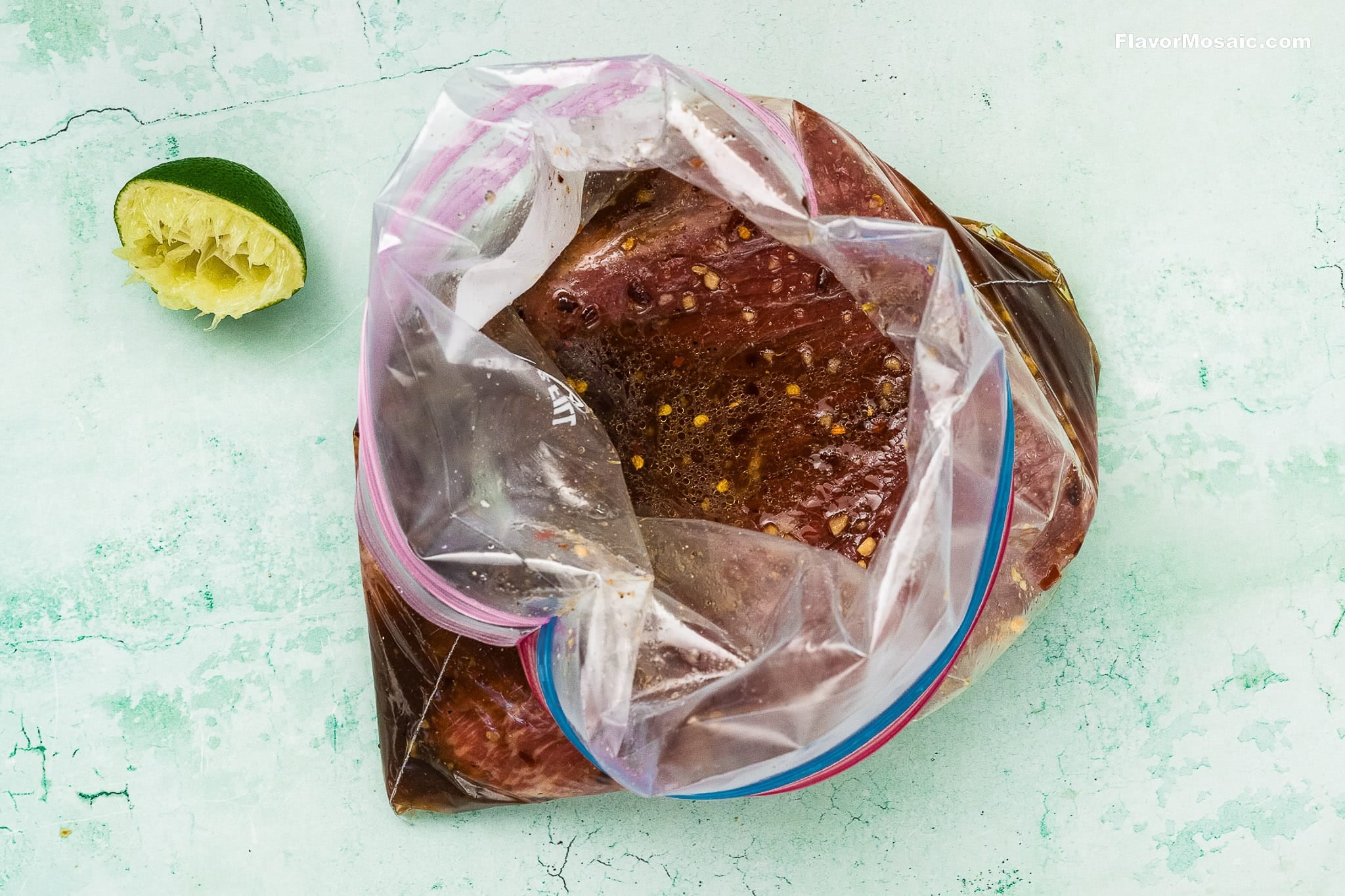 A plastic zip-top bag containing marinated Tex-Mex Beef Fajitas sits on a light green surface next to a squeezed lime half. The marinade appears to include spices and liquid seasoning.