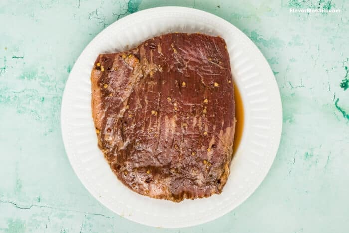 A marinated piece of raw brisket, perfect for Tex-Mex Beef Fajitas, sits on a white plate against a light green, textured background.