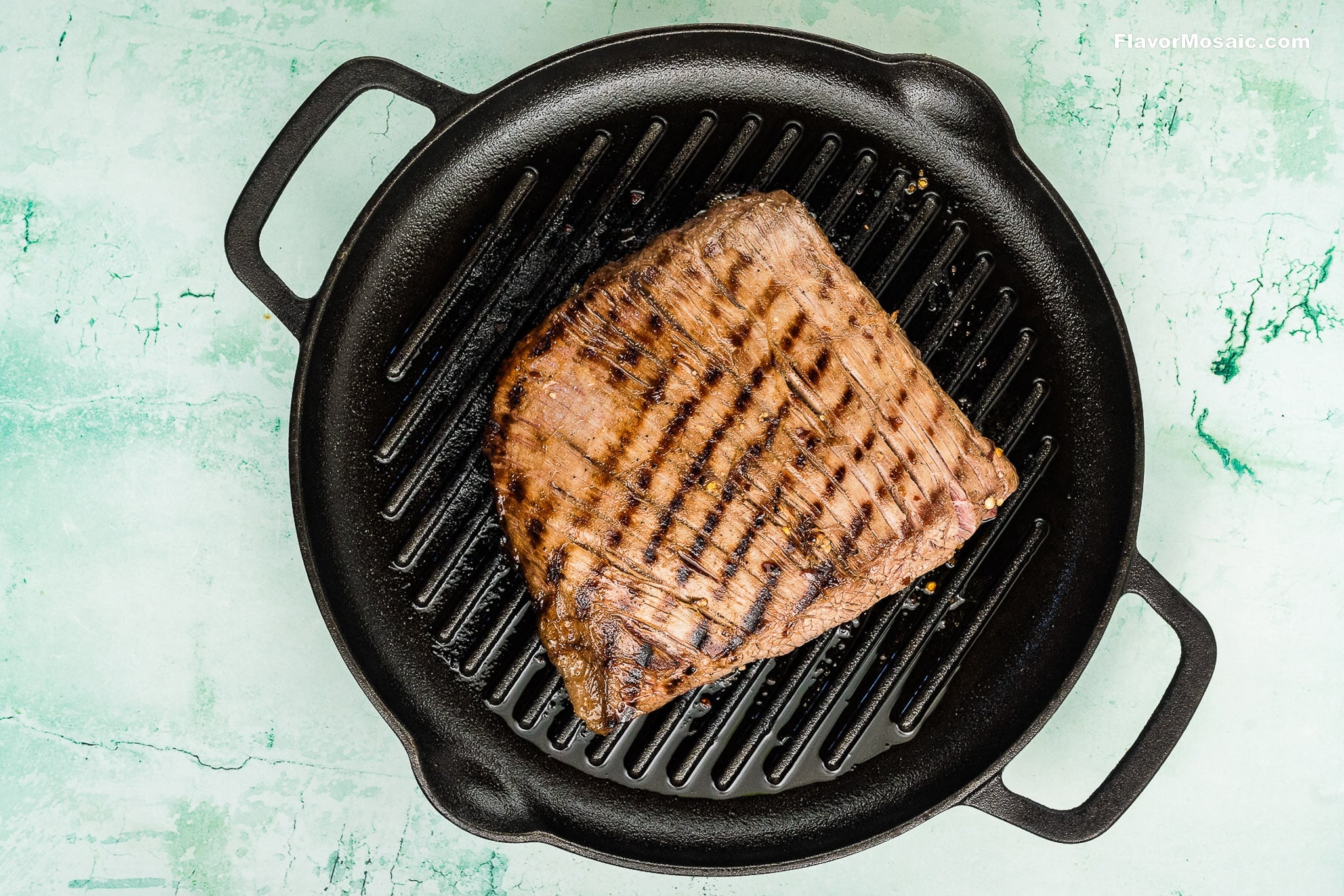 A grilled steak with visible sear marks sits on a black cast-iron grill pan, ready to be sliced for Tex-Mex Beef Fajitas, all placed on a light green, textured surface.