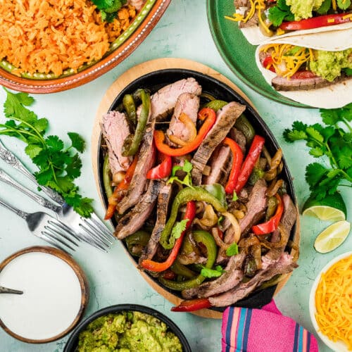 A colorful spread of Tex-Mex Beef Fajitas with grilled peppers and onions on a platter, served with tacos, guacamole, shredded cheese, refried beans, Mexican rice, and garnished with fresh cilantro and lime wedges.