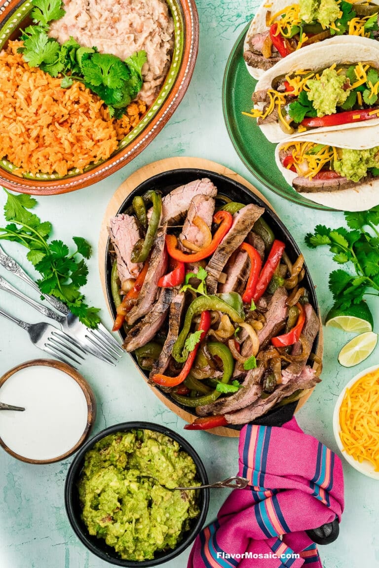 A colorful spread of Tex-Mex Beef Fajitas with grilled peppers and onions on a platter, served with tacos, guacamole, shredded cheese, refried beans, Mexican rice, and garnished with fresh cilantro and lime wedges.
