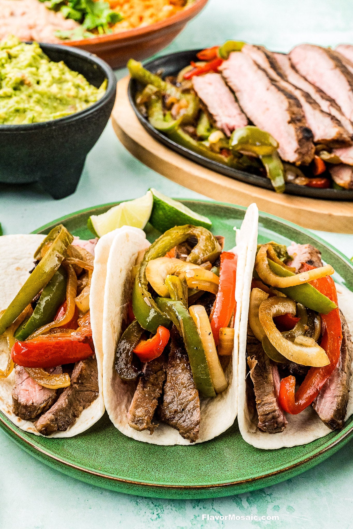 Three Tex-Mex Beef Fajitas tacos with sautéed bell peppers and onions on a green plate, served with lime wedges. In the background are sliced steak, guacamole, and a dish of rice.