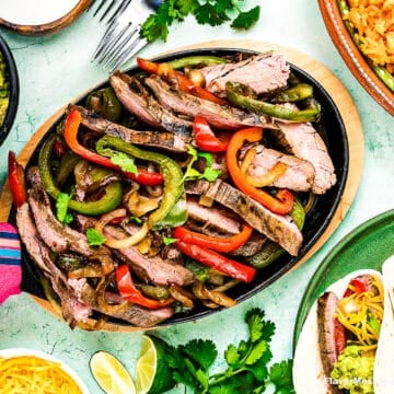 A platter of Tex-Mex Beef Steak Fajitas with grilled red and green bell peppers and onions is surrounded by guacamole, tacos, rice, lime wedges, and fresh cilantro. A hand holding a fork is reaching toward the sizzling fajitas.