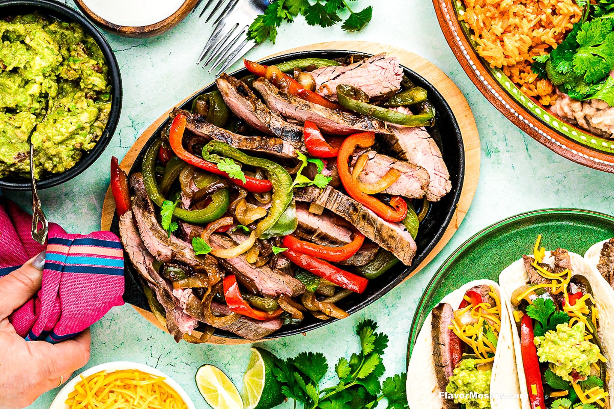 A platter of Tex-Mex Beef Fajitas with grilled red and green bell peppers and onions is surrounded by guacamole, tacos, rice, lime wedges, and fresh cilantro. A hand holding a fork is reaching toward the sizzling fajitas.