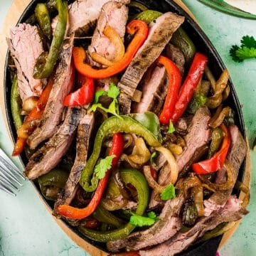 A sizzling platter of Tex-Mex Beef Fajitas with grilled steak strips, red and green bell peppers, and onions, garnished with cilantro, served on a black oval dish with a side of guacamole and tacos nearby.