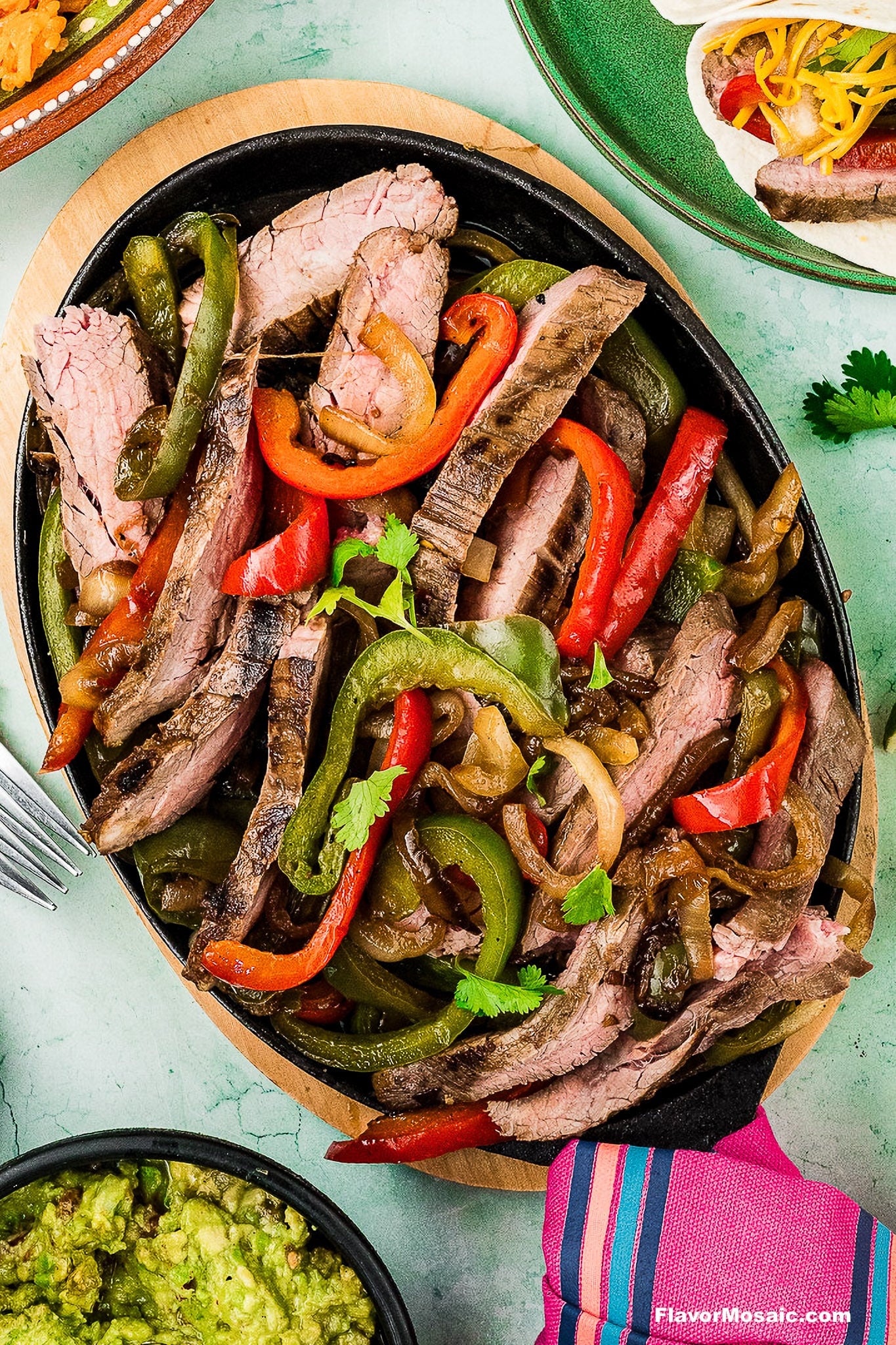 A sizzling platter of Tex-Mex Beef Fajitas with grilled steak strips, red and green bell peppers, and onions, garnished with cilantro, served on a black oval dish with a side of guacamole and tacos nearby.