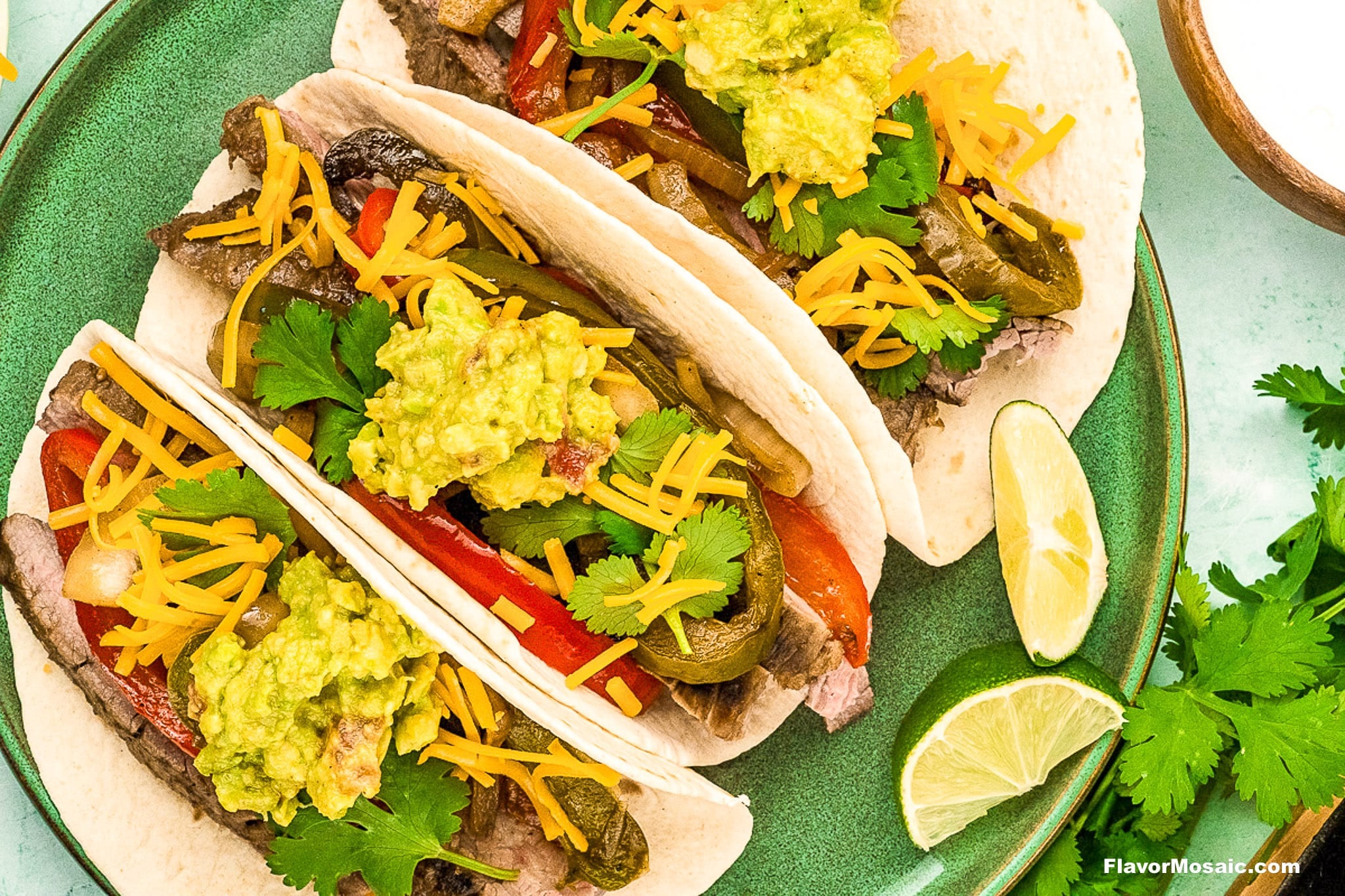 Three soft flour tortillas filled with sliced Tex-Mex beef fajitas, sautéed bell peppers, shredded cheddar cheese, guacamole, and fresh cilantro, served on a green plate with lime wedges and cilantro garnish.