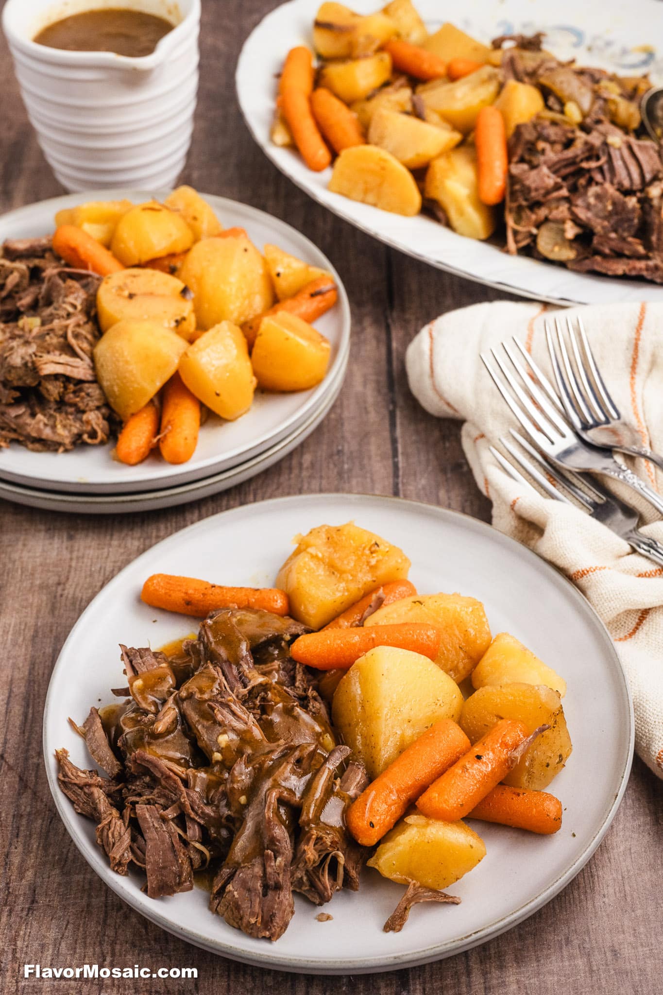 Two plates of Crock Pot Chuck Roast with shredded beef, carrots, and potatoes sit on a wooden table. In the background are a large serving dish filled with more food, a stack of sauce dishes, and a cloth napkin with forks.