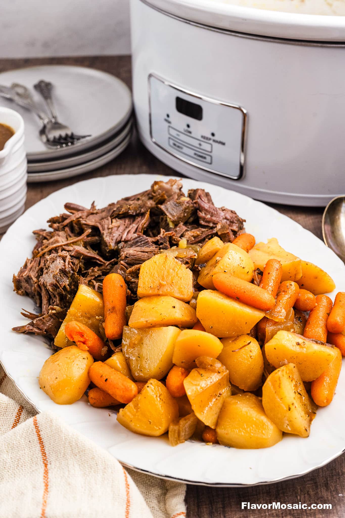 A white plate holds tender Crock Pot Chuck Roast, shredded with carrots and potatoes, in front of a slow cooker. Nearby, stacked plates, bowls, and forks rest on a wooden table with a striped napkin.