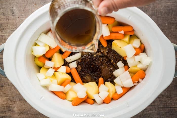 A hand pours brown broth from a glass measuring cup into a slow cooker filled with chunks of potatoes, carrots, onions, and seasoned meat, preparing a hearty Crock Pot Chuck Roast.