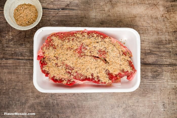 Raw steak in a white foam tray topped with a dry spice rub, perfect for Crock Pot Chuck Roast, sits on a rustic wooden surface. A small bowl of extra spice mixture is to the top left. FlavorMosaic.com appears in the bottom left corner.