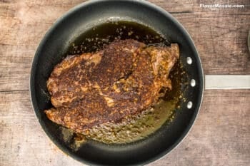 A large, seasoned steak reminiscent of a Crock Pot Chuck Roast sizzles in a nonstick skillet with rich juices and oil, resting on a rustic wooden surface.