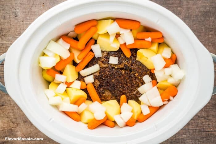 A white Crock Pot contains a chuck roast in the center, surrounded by neatly arranged, uncooked potatoes, carrots, and onions-perfect for making a classic Crock Pot Chuck Roast.