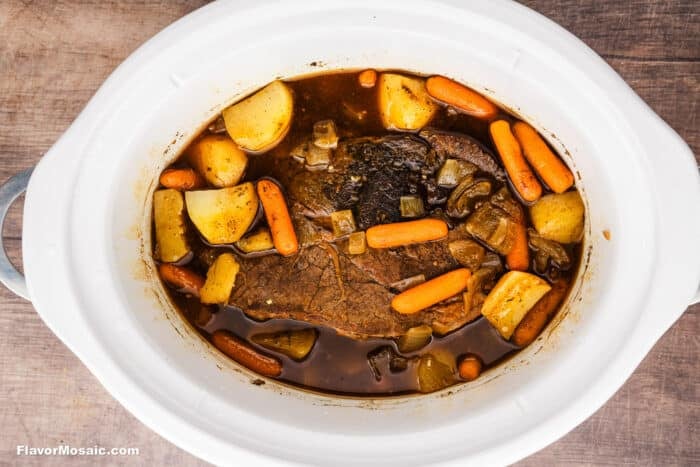 A Crock Pot Chuck Roast simmers in a slow cooker, surrounded by baby carrots, potato chunks, onion pieces, and rich brown broth on a wooden surface.