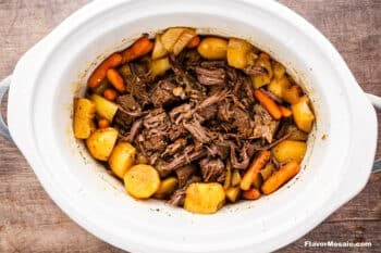 A cooked Crock Pot Chuck Roast with shredded beef, carrots, potatoes, and onions in a white slow cooker, sitting on a wooden surface.