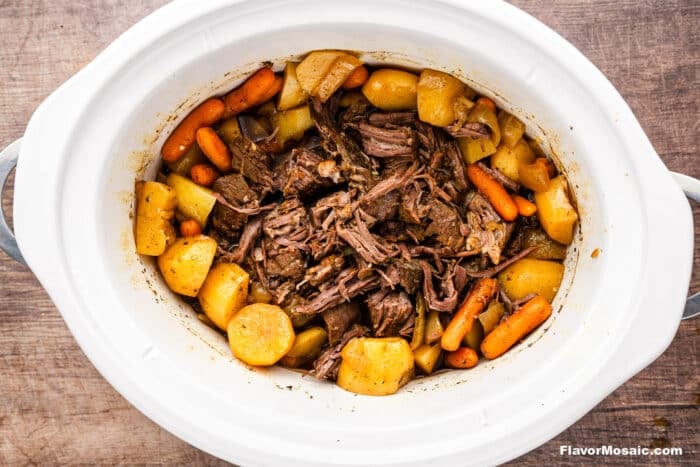 A white slow cooker filled with Crock Pot Chuck Roast, surrounded by cooked carrots, potatoes, and onions, all in a rich broth on a wooden surface.