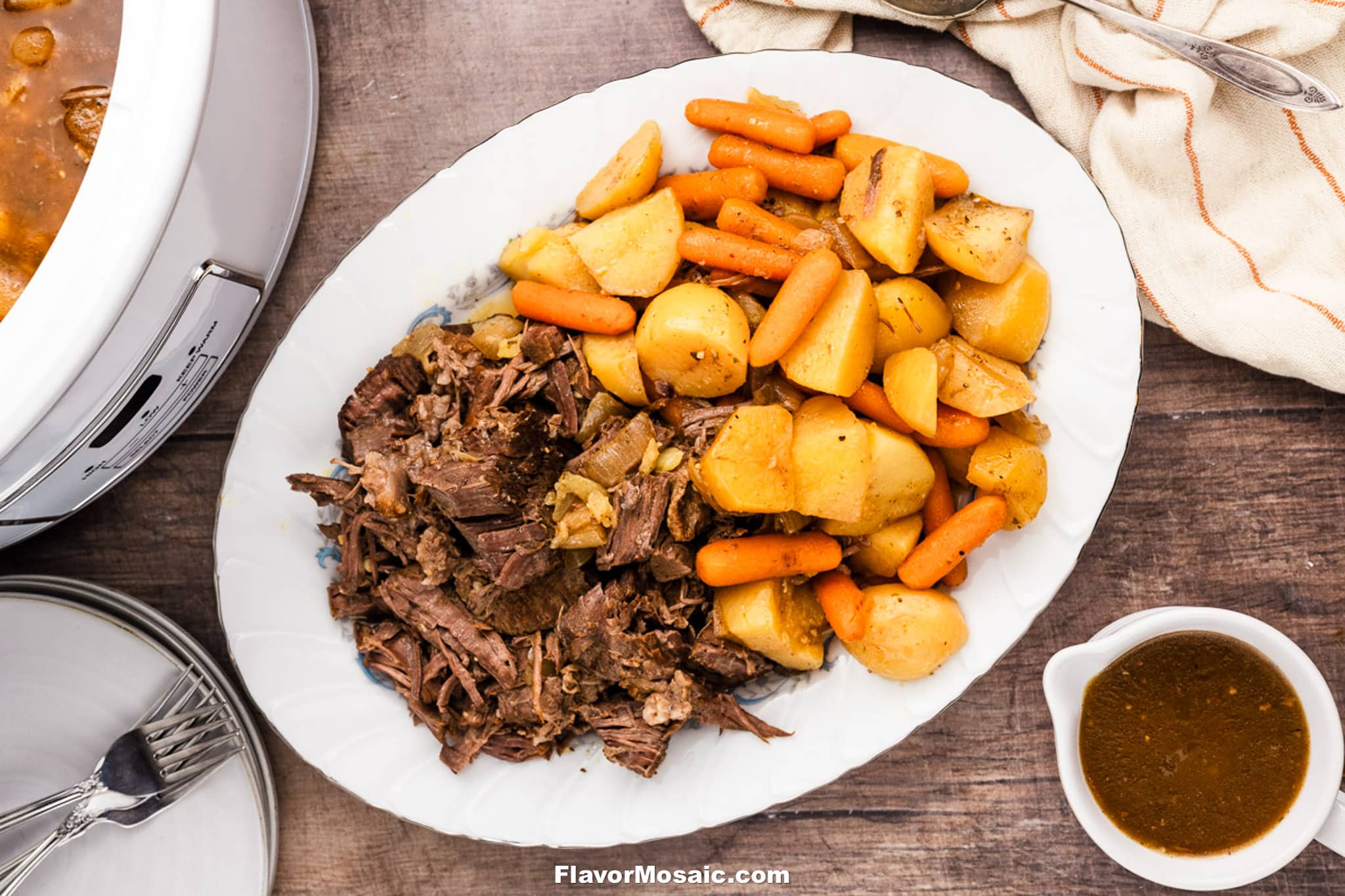An overhead view of a platter with Crock Pot Chuck Roast, shredded pot roast, cooked potatoes, and baby carrots. A small bowl of gravy, plates, and a slow cooker are nearby on a wooden table.