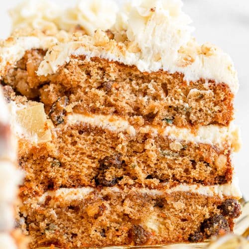 Close up view of the side of a slice of 3-layer homemade carrot cake with cream cheese frosting, and white marble background.