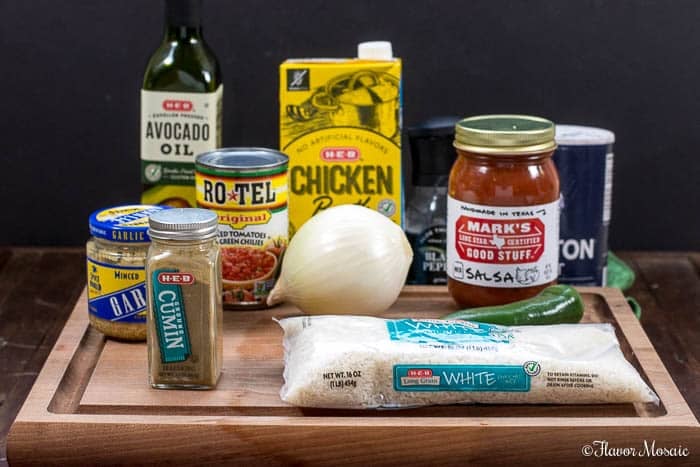 A wooden cutting board holds the ingredients for Mexican Rice—white rice, white onion, jalapeño, can of Rotel, minced garlic, ground cumin, salsa, avocado oil, chicken broth, and salt—all arranged upright against a dark background.