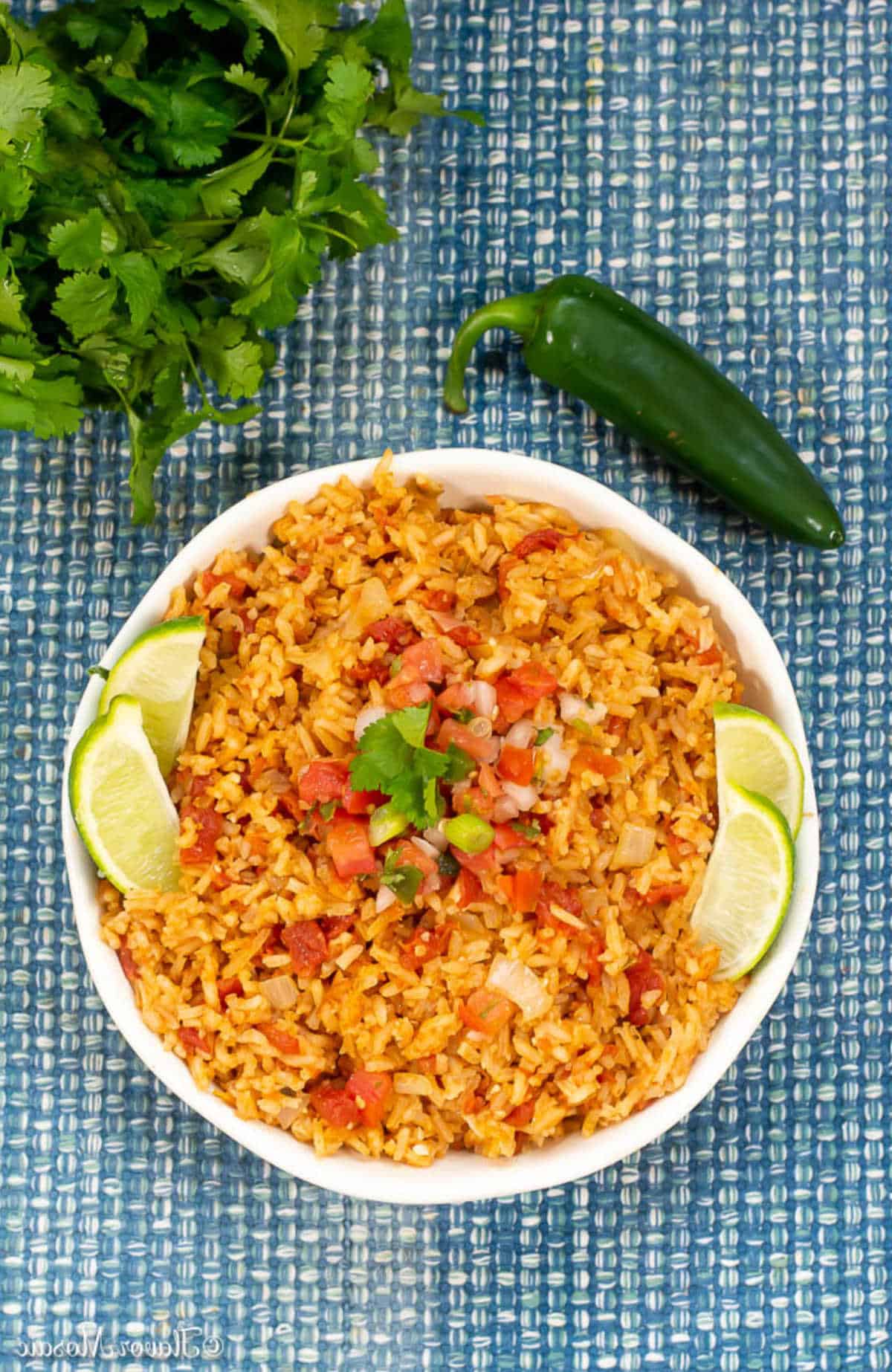 A bowl of flavorful Mexican Rice garnished with lime wedges and cilantro, surrounded by fresh cilantro and a whole jalapeño pepper, set on a blue woven placemat.