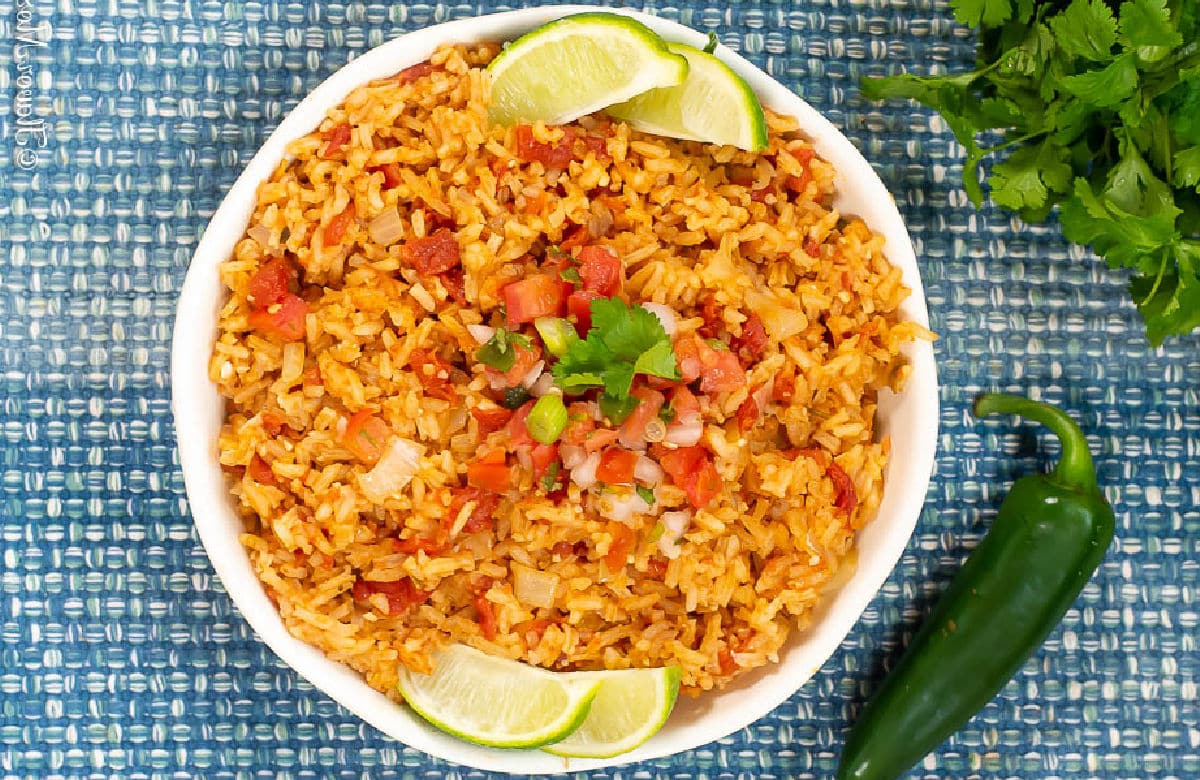 A bowl of Mexican Rice topped with chopped tomatoes and cilantro, garnished with lime wedges. A jalapeño pepper and fresh cilantro are beside the bowl on a blue woven placemat.