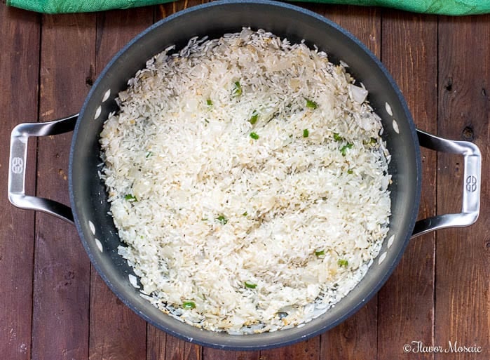 Overhead view of a pot filled with uncooked Mexican Rice mixed with chopped onions and green herbs, placed on a wooden surface.