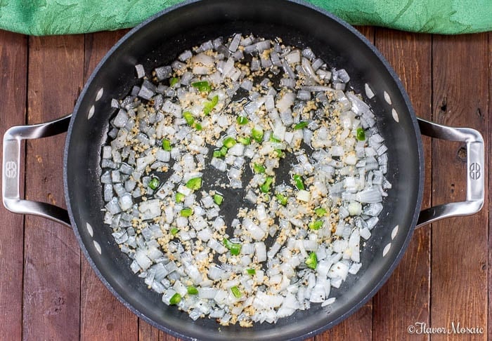 Chopped onions, garlic, and green peppers are sautéing in a large black skillet with metal handles, the perfect start for flavorful Mexican Rice, set on a wooden surface with a green cloth nearby.