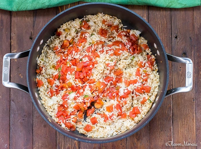 A large black pot of Mexican Rice, filled with cooked rice, diced tomatoes, and chopped onions, sits on a wooden surface, with a green cloth partially visible at the top.