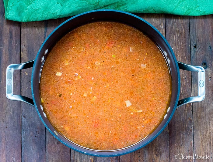 A pot of tomato-based vegetable soup with visible diced vegetables sits on a wooden surface, viewed from above. A green cloth is partially visible at the top, evoking the vibrant colors of Mexican Rice.