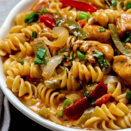 White bowl with Chicken Fajita Pasta with red and green bell peppers and garnished with chopped green onions and cilantro with a gray napkin on dar, wood table.