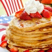 Stack of Strawberry Pancakes topped with strawberries, maple syrup, and whipped cream, on white plate with bowl of strawberries in the background.