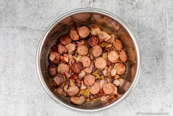 A metal pot filled with sliced, browned sausage and bacon pieces sits on a light gray, textured countertop-perfect ingredients to start flavorful Instant Pot Black Eyed Peas.