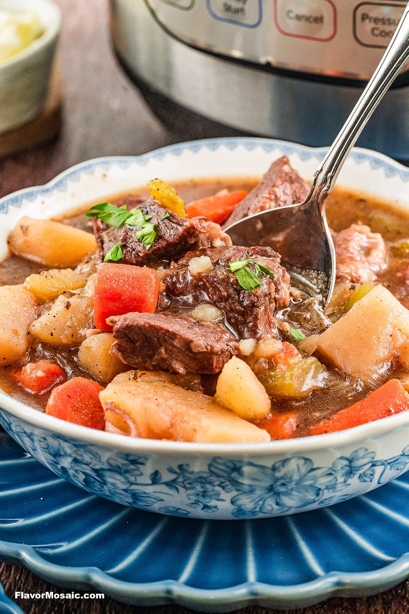 A bowl of hearty Instant Pot Beef Stew with chunks of beef, carrots, and potatoes, garnished with fresh parsley, sits on a blue-patterned plate as a spoon lifts a bite; a pressure cooker is in the background.