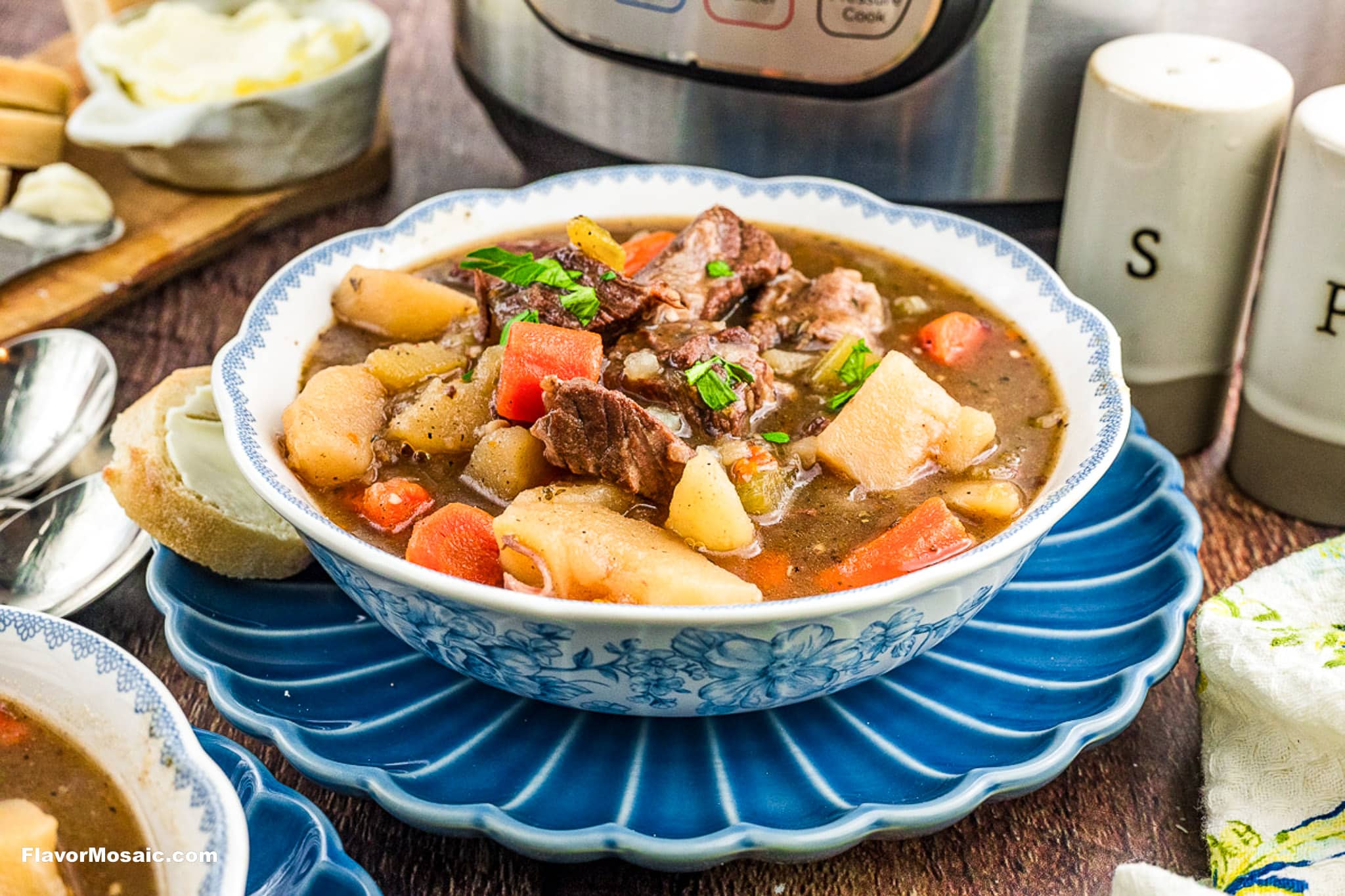 A bowl of Instant Pot Beef Stew with chunks of beef, potatoes, carrots, and peas, garnished with herbs, sits on a blue plate next to a slice of bread, salt and pepper shakers, and a slow cooker in the background.